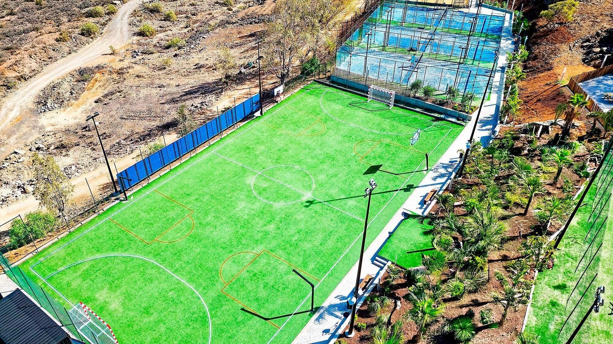 Vista aérea de un campo deportivo con una valla perimetral, un camino de tierra y vegetación escasa.