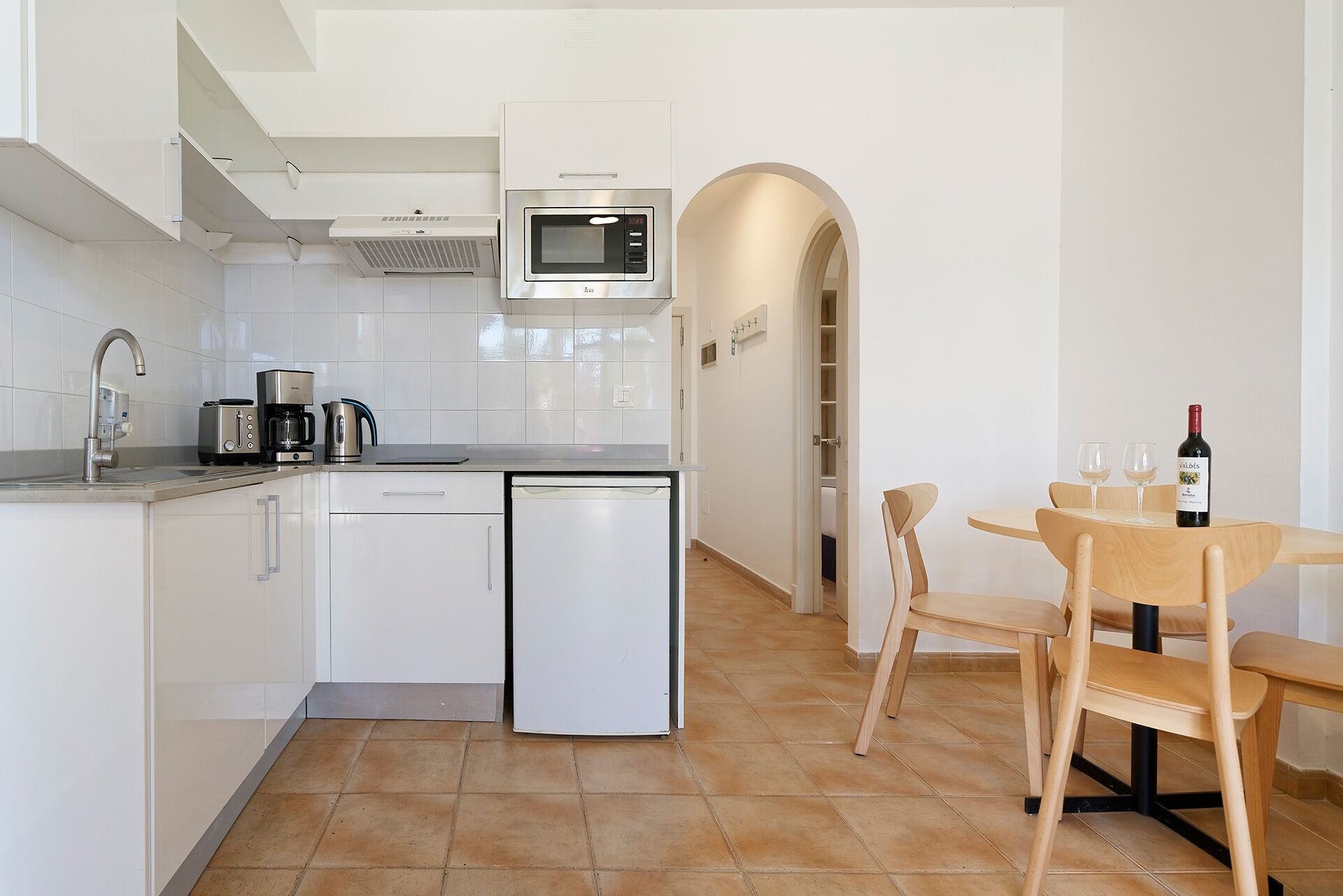A modern kitchen with white cabinets, a built-in microwave, and a dishwasher. There's a dining area with a wooden table and chairs, and a wine bottle on the table.
