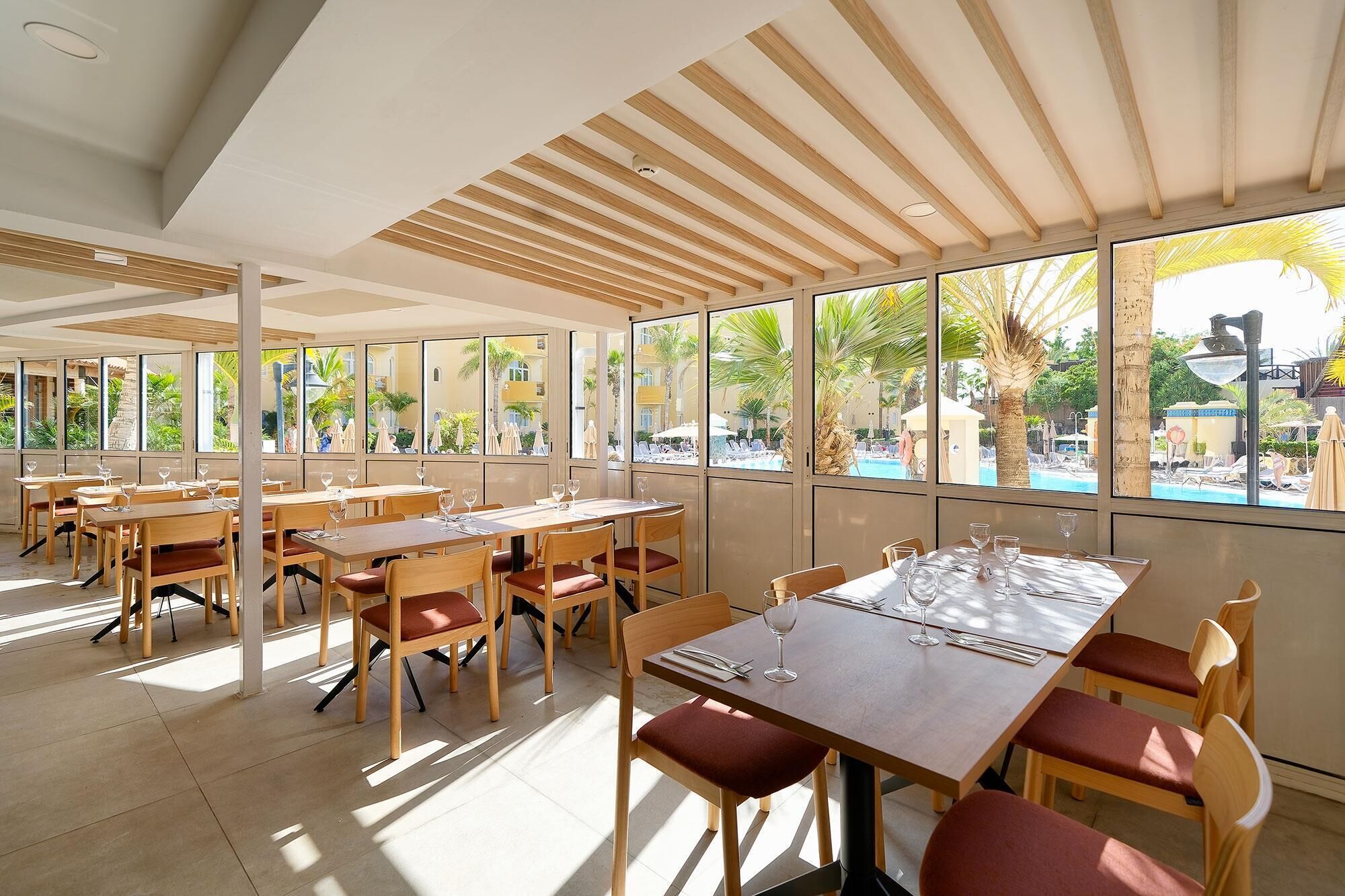 A restaurant with wooden tables and chairs, large windows with a view of a pool and palm trees.