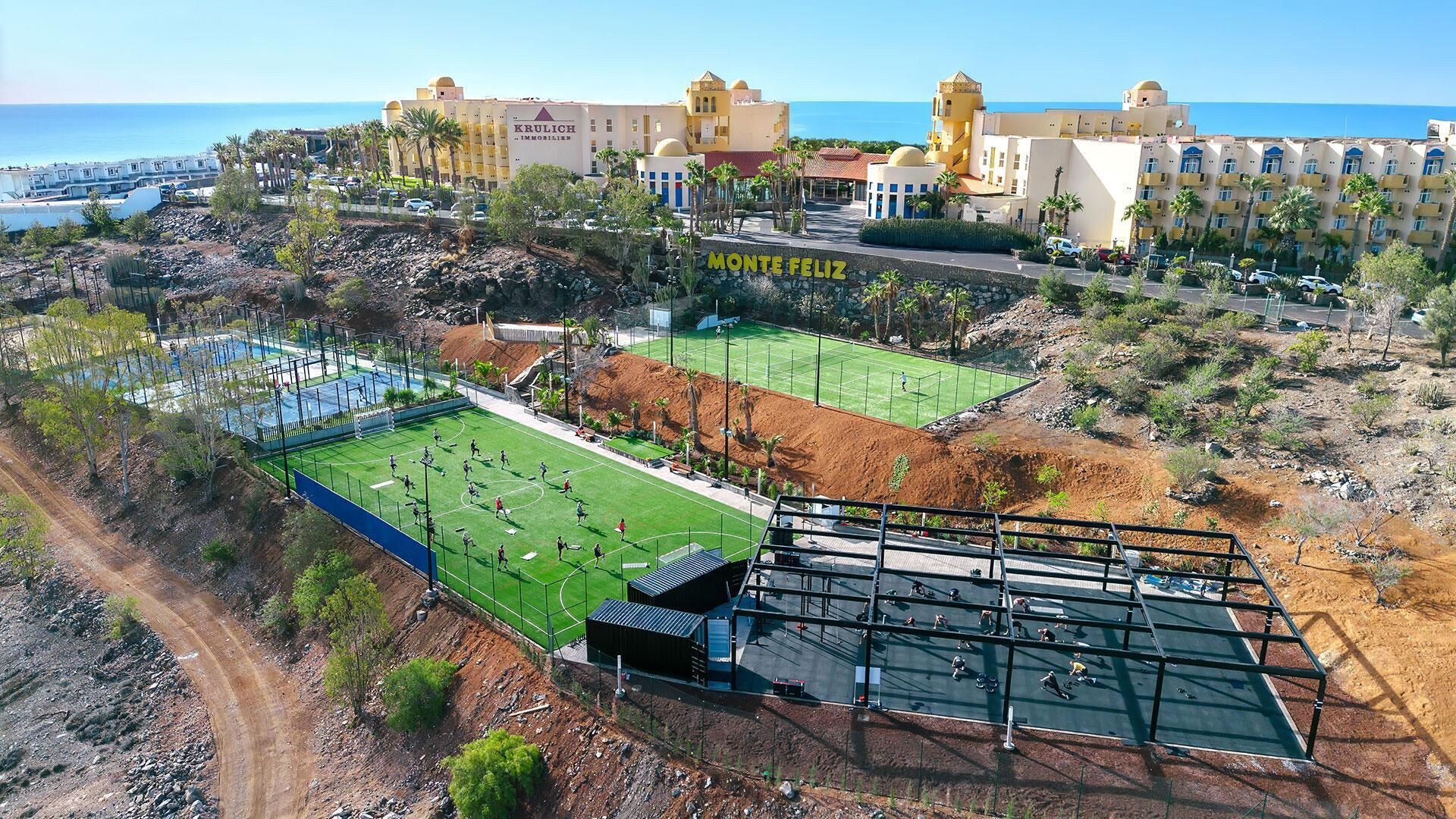 A coastal hotel complex with multiple buildings, a sports field, and a parking area.
