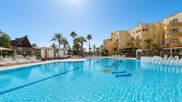 A swimming pool with lounge chairs and umbrellas, surrounded by palm trees and hotel buildings. - Sport Hotel Monte Feliz - powered by Playitas (San Bartolomé de Tirajana)
