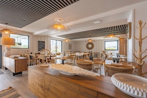 Un espace repas moderne avec des tables et des chaises en bois, des luminaires suspendus et un îlot central agrémenté d’un élément décoratif blanc.
