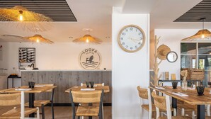 A restaurant interior with wooden tables and chairs, woven ceiling lamps, a wall clock, and a logo on the wall. - Relais Thalasso île de Ré - Hôtel Atalante (Sainte-Marie-de-Re)