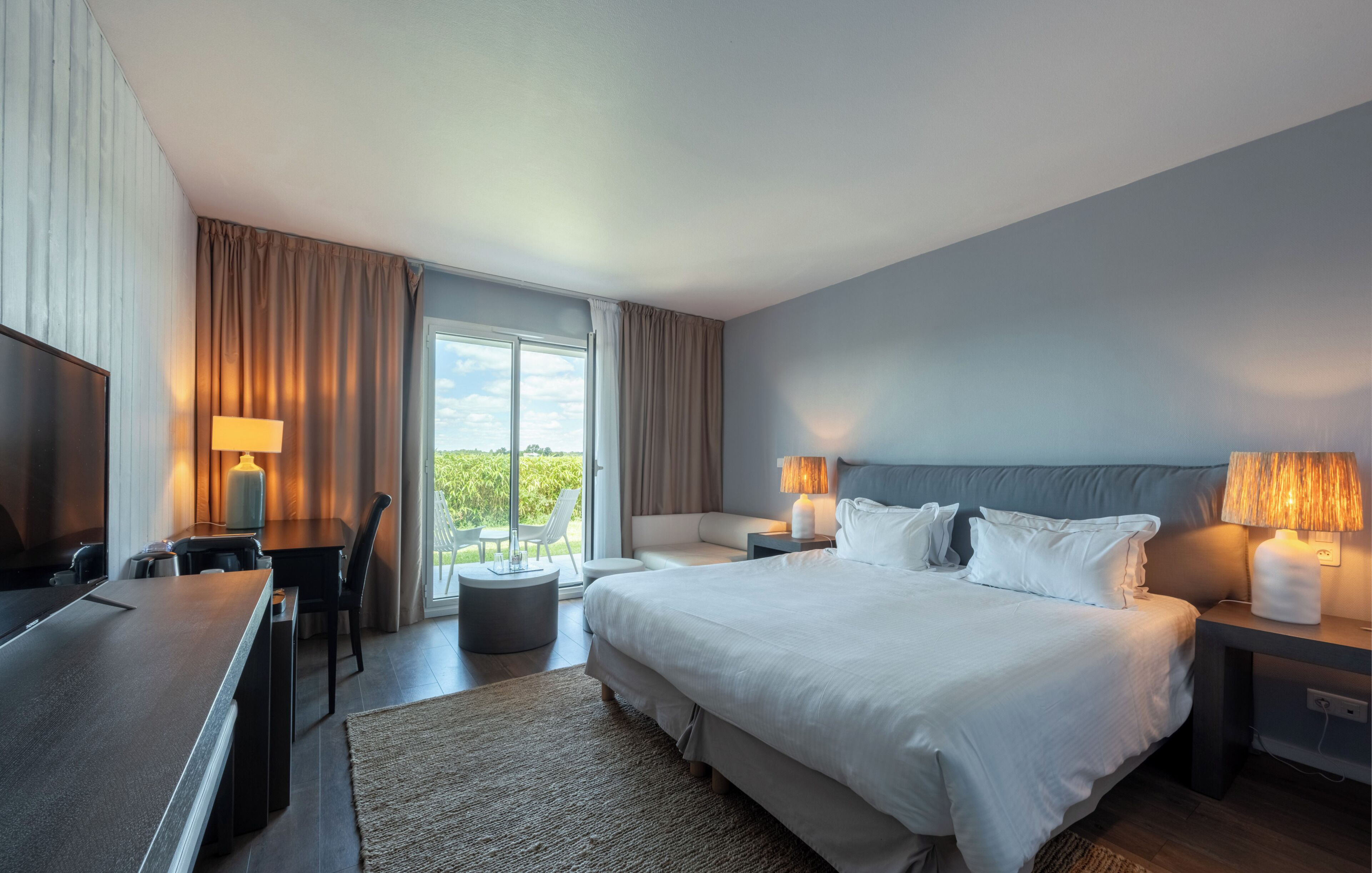 Een hotelkamer met een bed, bedlampen, een bureau met lamp, een televisie en uitzicht op de omgeving.