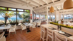 Un espace de restauration intérieur avec des tables et des chaises blanches, de grandes fenêtres donnant sur la mer et un ventilateur de plafond.