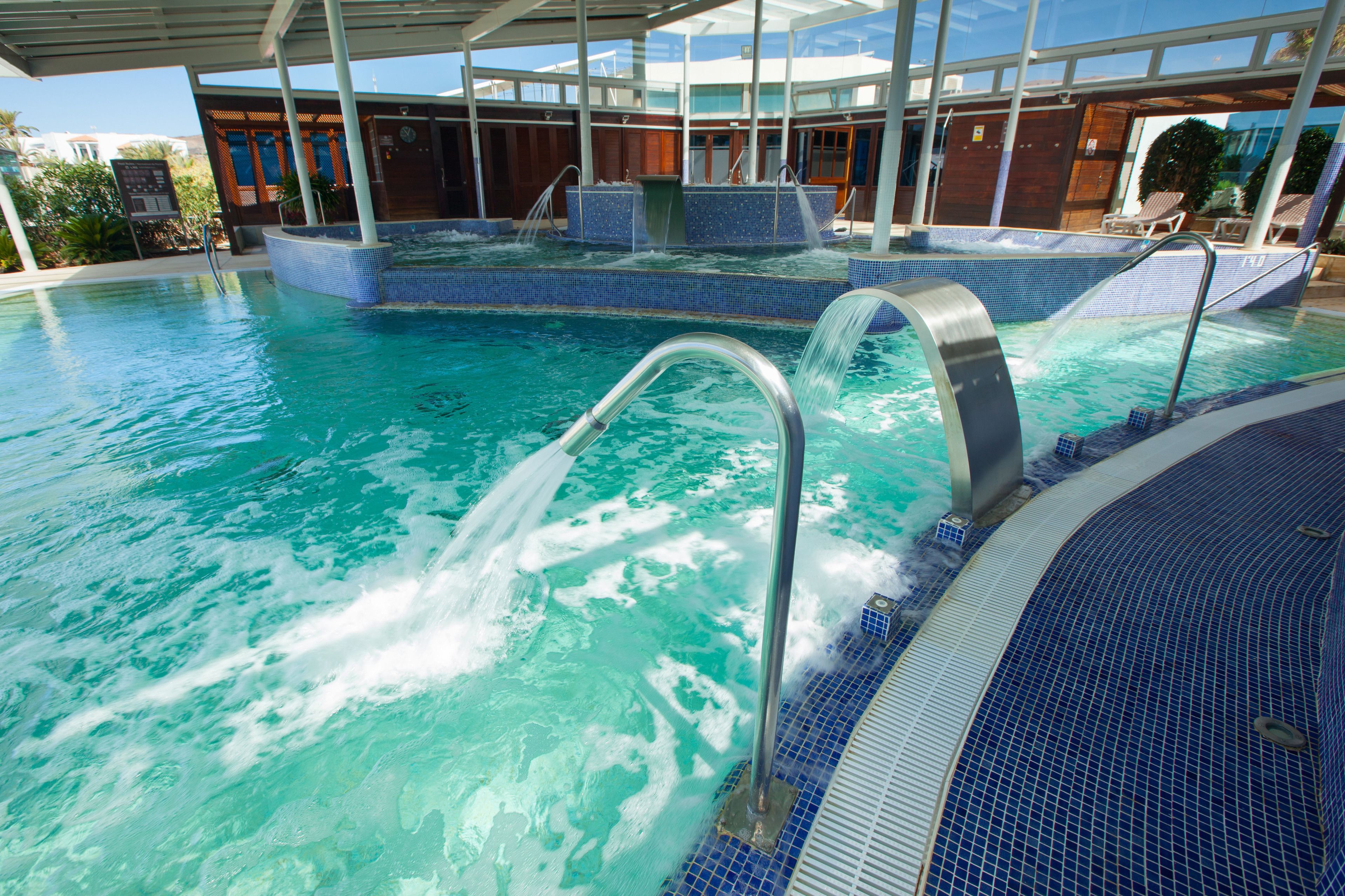 Una gran piscina interior con agua cristalina, zona de jacuzzi y área de descanso cubierta con sillones reclinables.