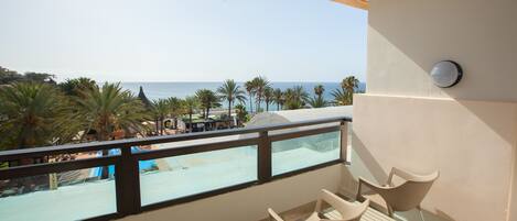 A balcony with a view of the beach and palm trees. There are two chairs and a table on the balcony.