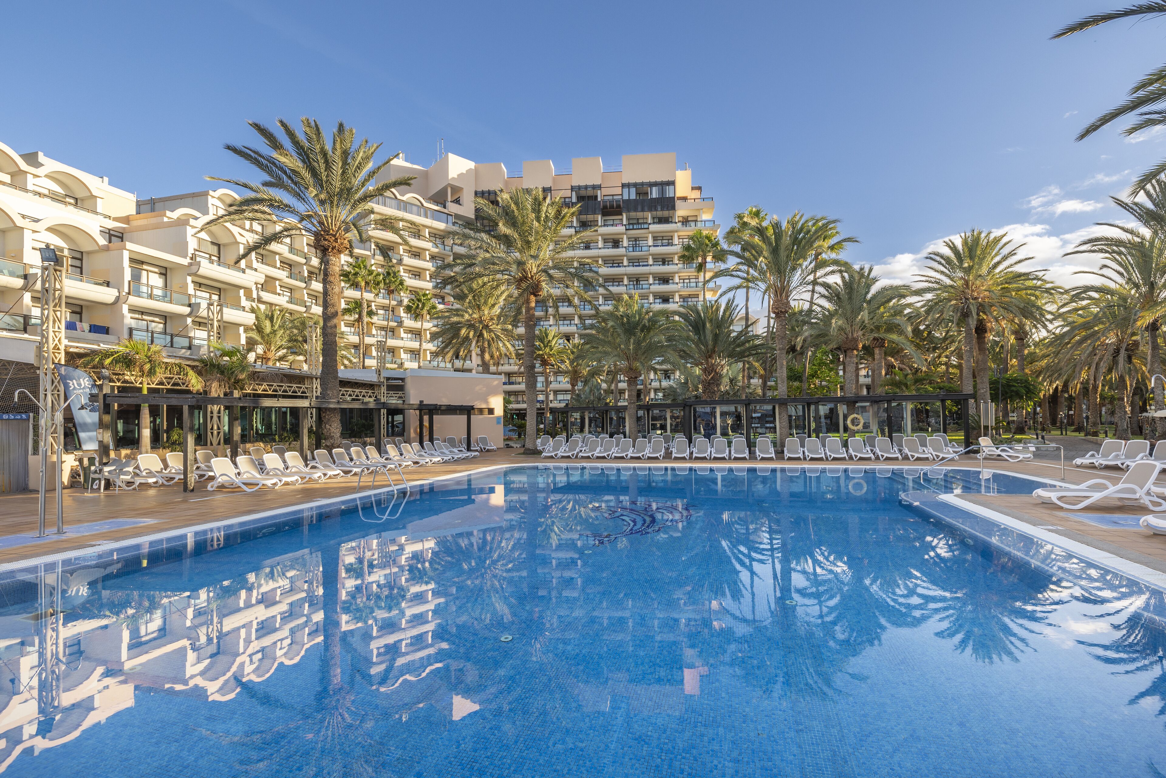 A swimming pool with lounge chairs, palm trees, and a multi-story hotel building in the background.