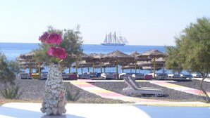 Ein Strandresort mit Liegestühlen und Sonnenschirmen, einer Steinsäule mit Blumen und einem Segelboot im Hintergrund.