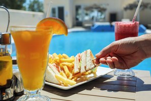 Une table à manger avec une assiette de nourriture, une boisson garnie d’une tranche d’orange, et une piscine en arrière-plan.