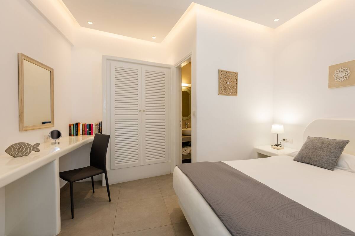 A bedroom with a bed, bedside table, lamp, chair, desk, mirror, and wardrobe.