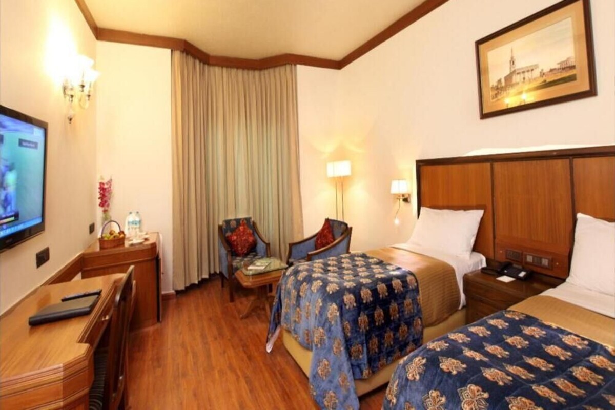 A hotel room with two beds, a desk, a TV, a lamp, and a painting on the wall.
