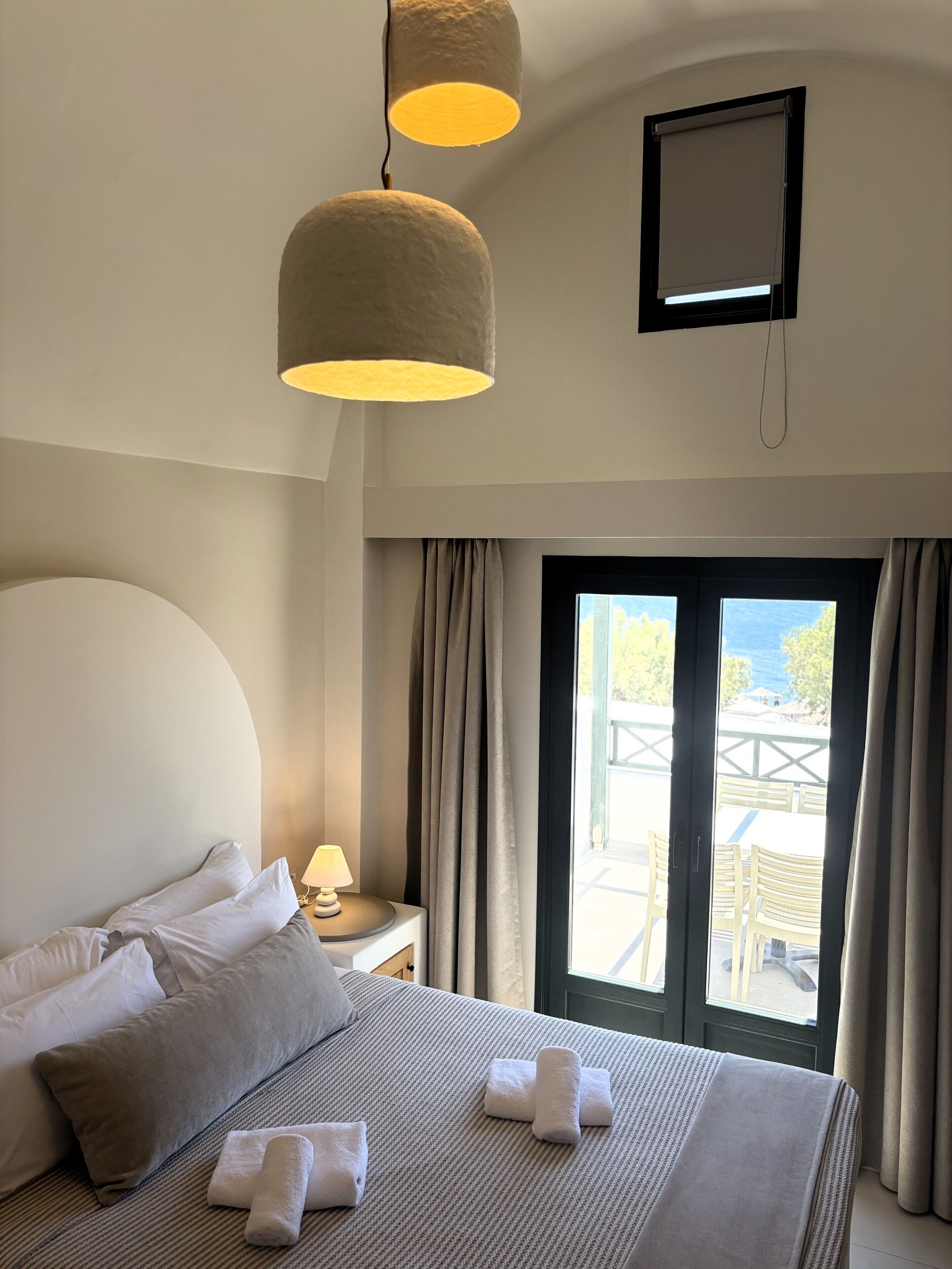 A hotel room with a bed, a bedside table with a lamp, a window with curtains, and a view of the outside.