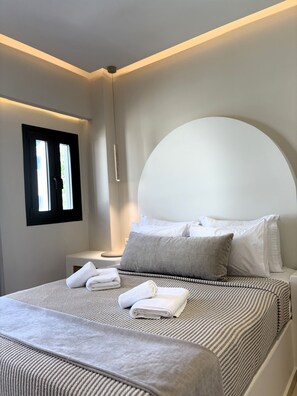 A modern bedroom with a large bed, a bedside table, a window, and a wall-mounted light fixture. - Sellada Beach  (Santorini)
