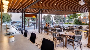 An outdoor cafe with wooden tables and chairs, a covered seating area, and a view of the sea.