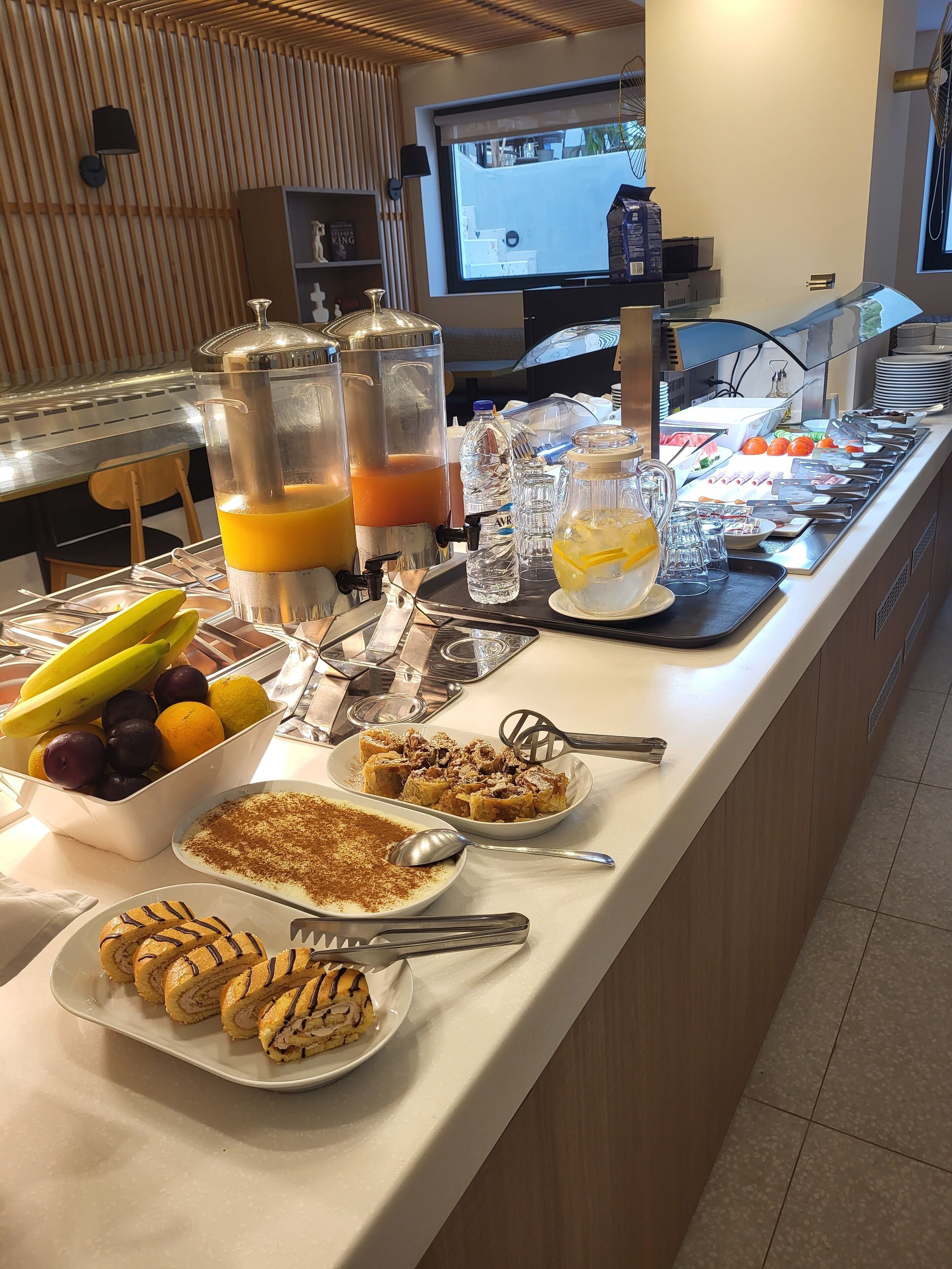 Ein reichhaltiges Buffet mit verschiedenen Speisen und Getränken, darunter Obst, Gebäck und ein großer Bildschirm im Hintergrund.