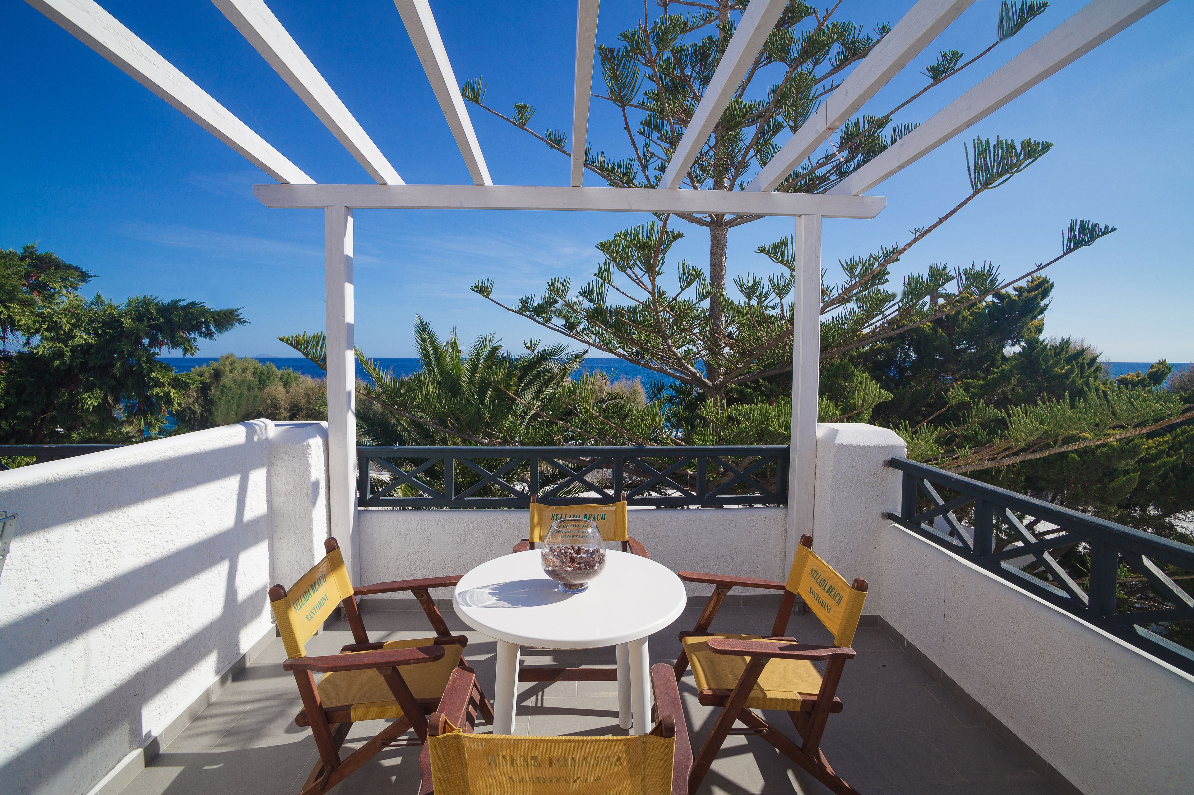 A covered outdoor seating area with a white table and wooden chairs, overlooking a scenic view of the sea and greenery.