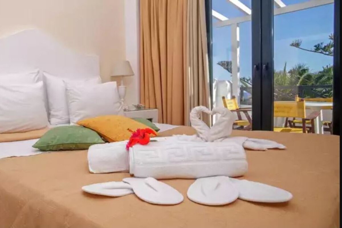A hotel room with a bed, pillows, a nightstand, a lamp, and a view of the outdoors.