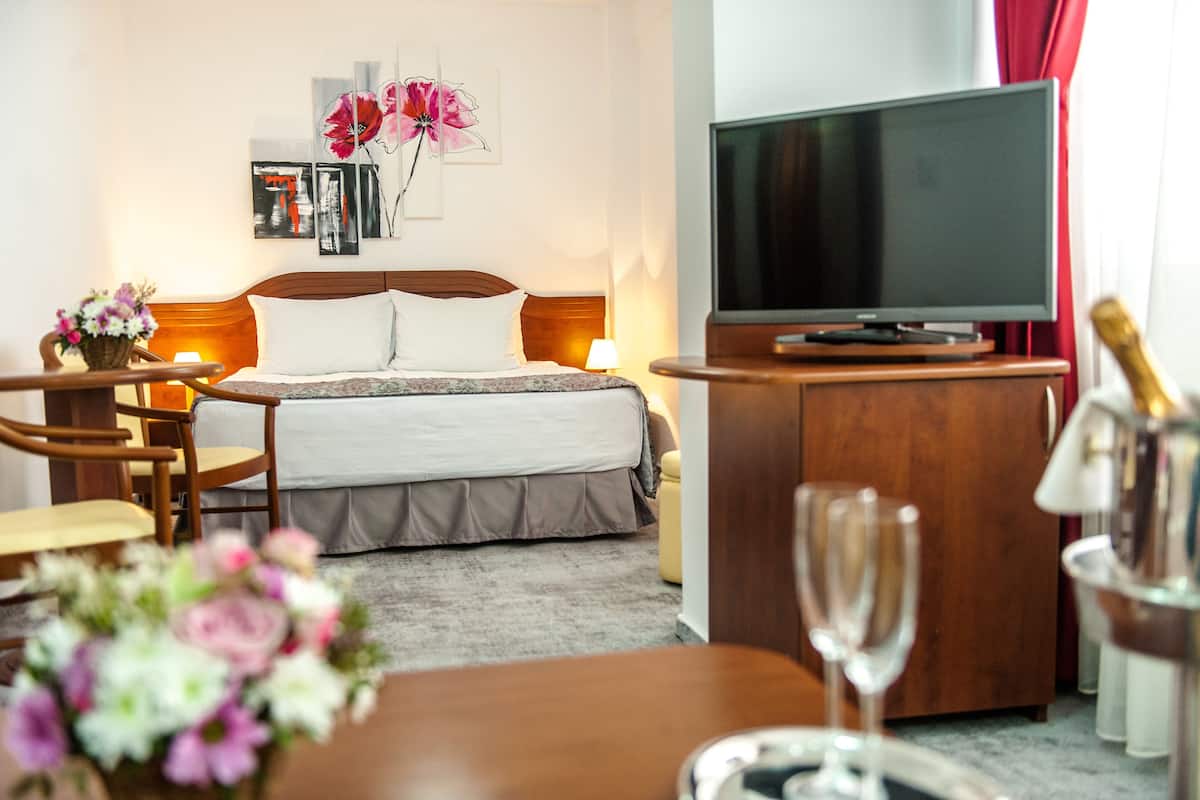 A hotel room with a bed, a television, a wooden desk, a chair, a vase of flowers, and a champagne glass.