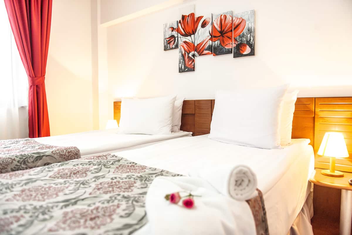 A hotel room with two beds, a wooden headboard, a wall-mounted artwork of red flowers, and a bedside lamp.