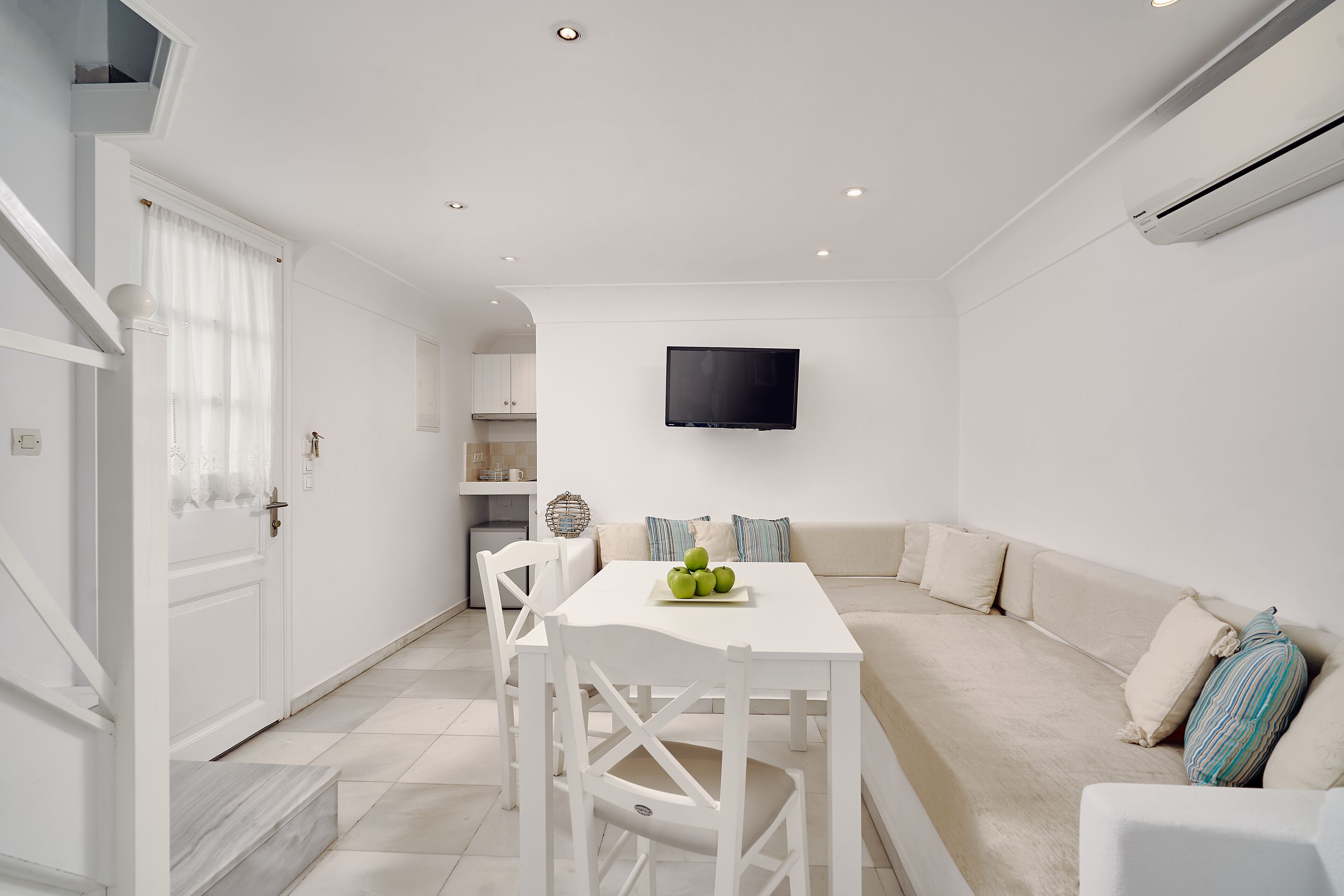 A modern, white-themed living room with a dining area, a flat-screen TV mounted on the wall, and a sofa with cushions.
