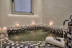 A bathroom with a tiled bathtub, candles, and a window. - Merovigliosso Apartments (Santorini)