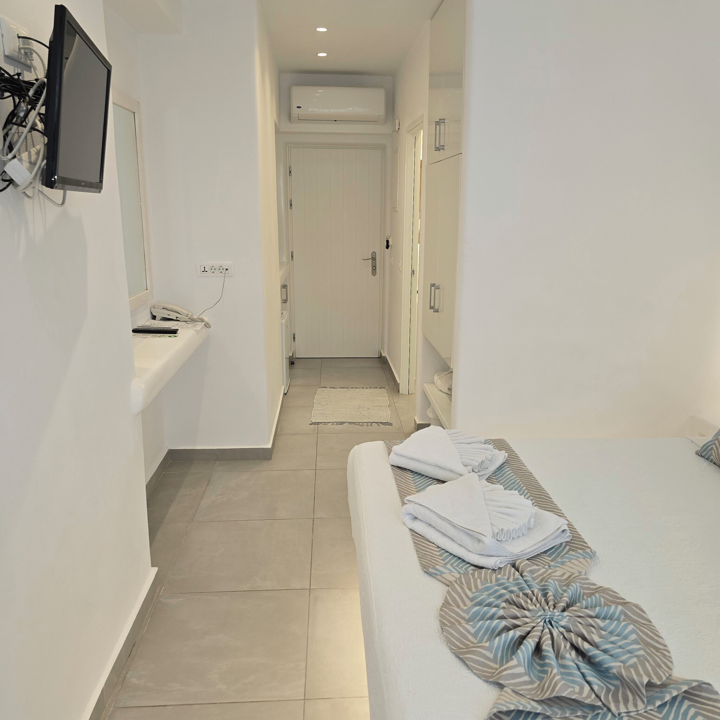 Habitación de hotel con cama, televisión montada en la pared, escritorio con teléfono y cargador, y equipo de aire acondicionado sobre la puerta.