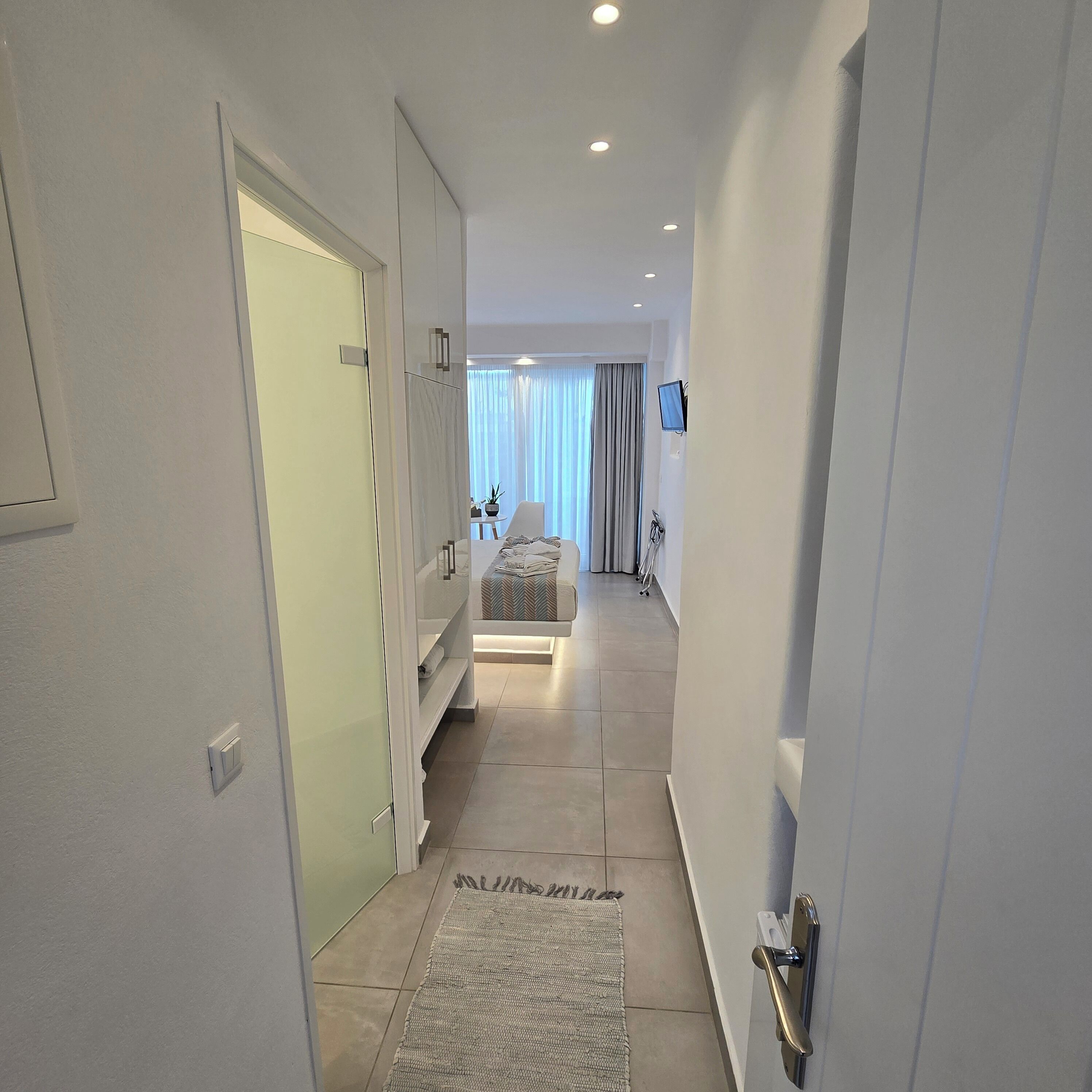 A hallway leading to a bedroom with a bed, a TV, and a window with curtains.