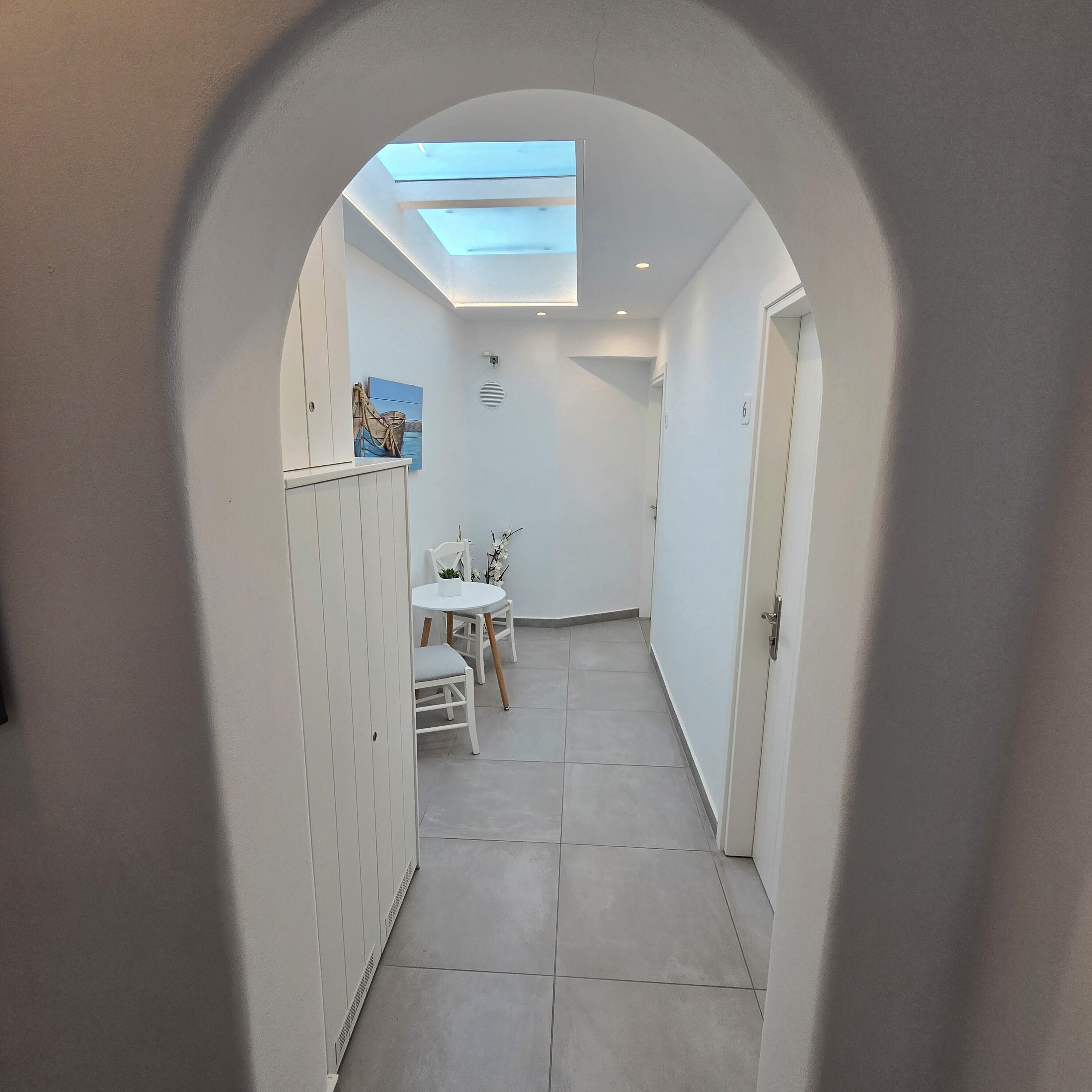A narrow hallway with a skylight, a small round table with chairs, and a cabinet with a painting.
