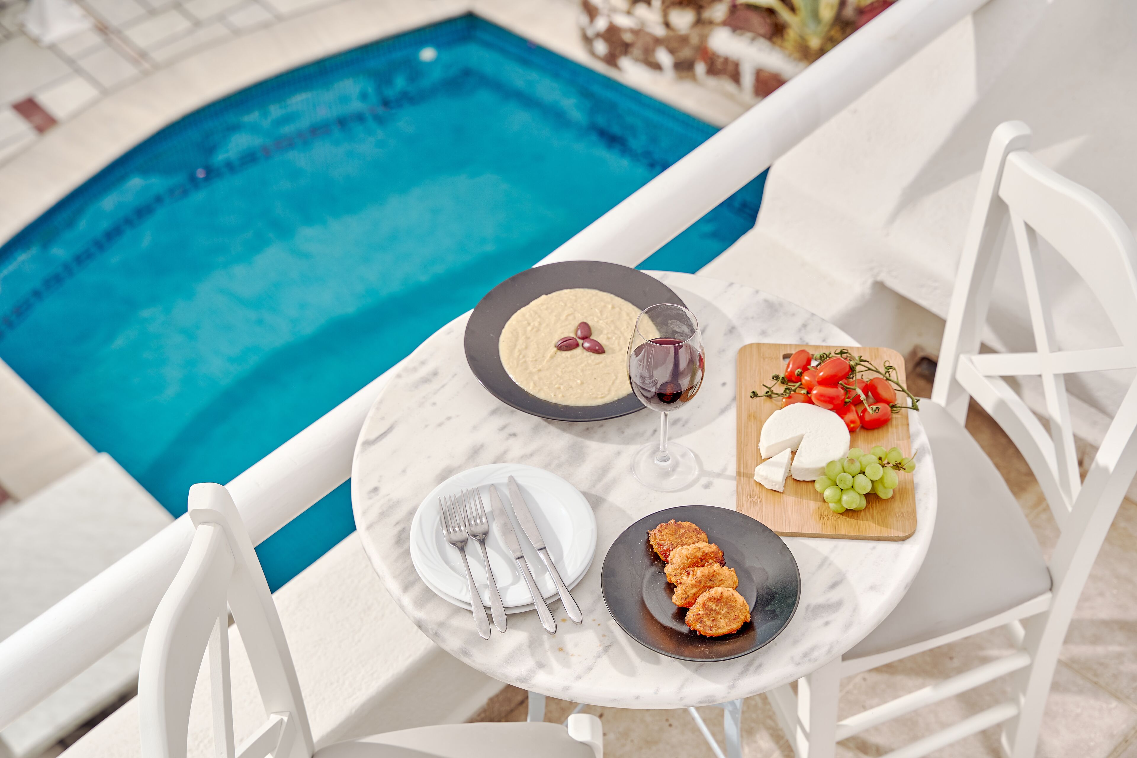 A dining table with plates, cutlery, and a glass of red wine, set against a poolside view.