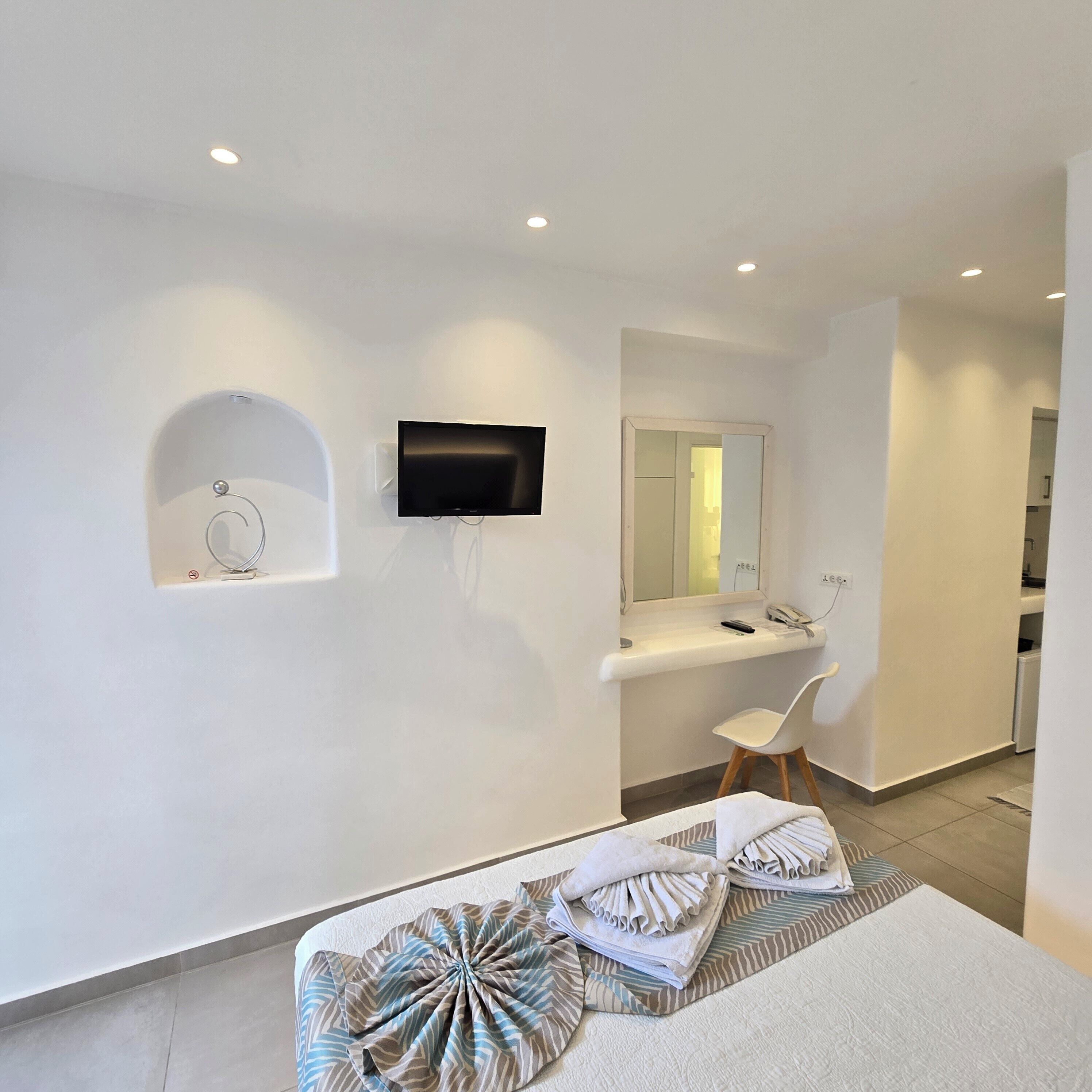 Una habitación de hotel moderna con televisión de pantalla plana montada en la pared, una tocador con espejo y una pequeña zona de estar con una silla.