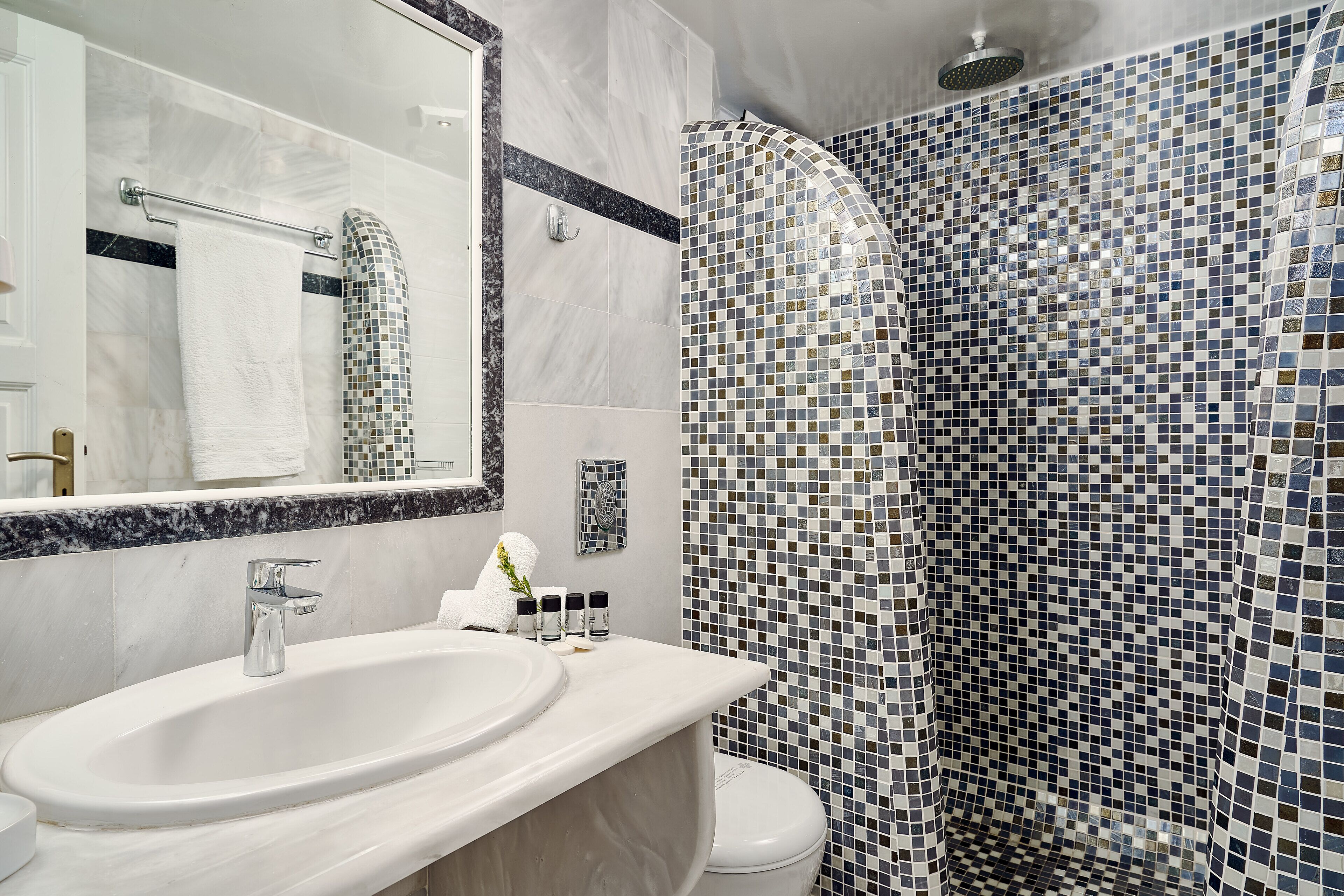 Un baño con lavamanos blanco, un espejo grande y una ducha con mosaicos.