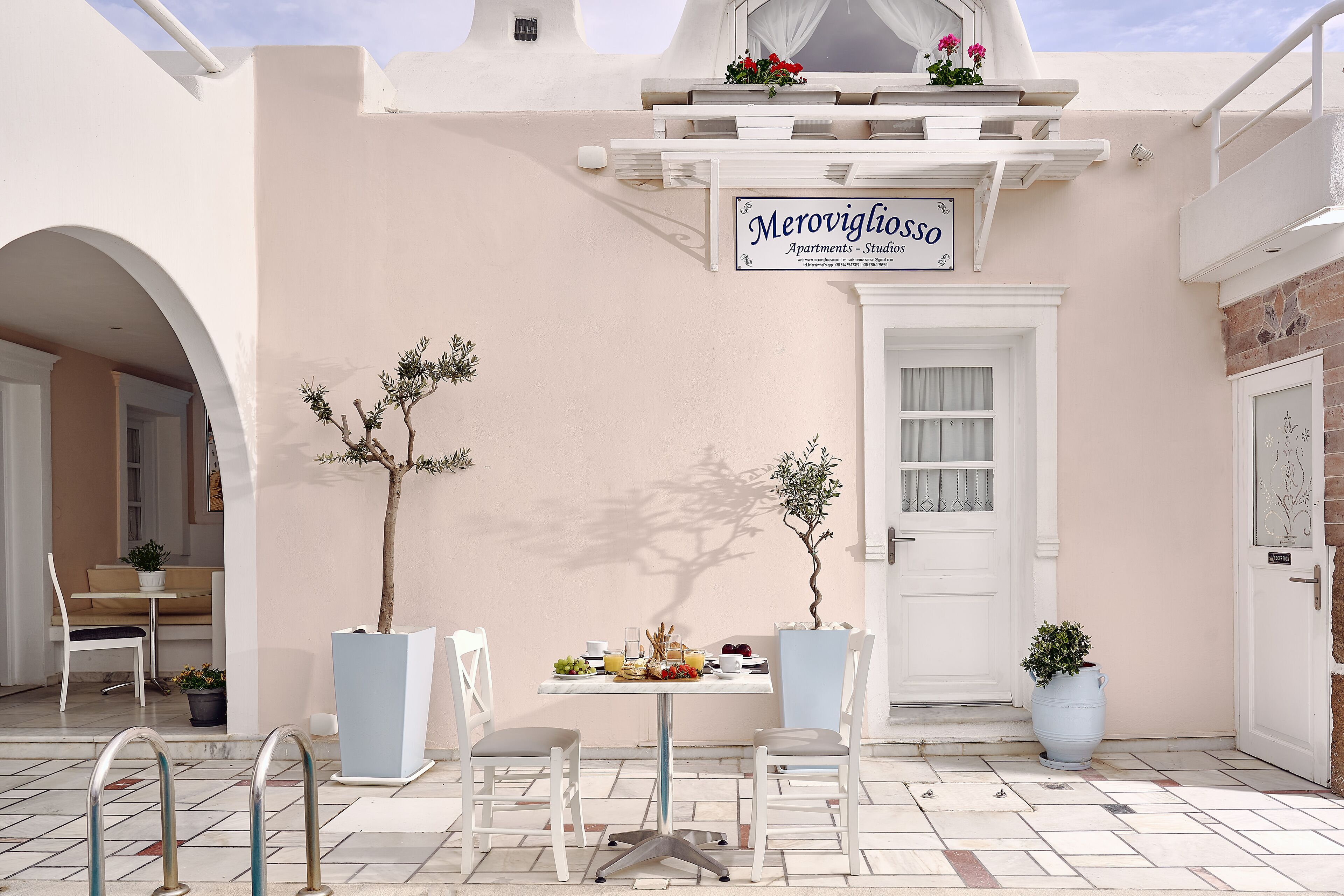A building with a sign reading "Merovigliasso Apartments - Studios." There is a table with food and drinks, two potted plants, and a white door.