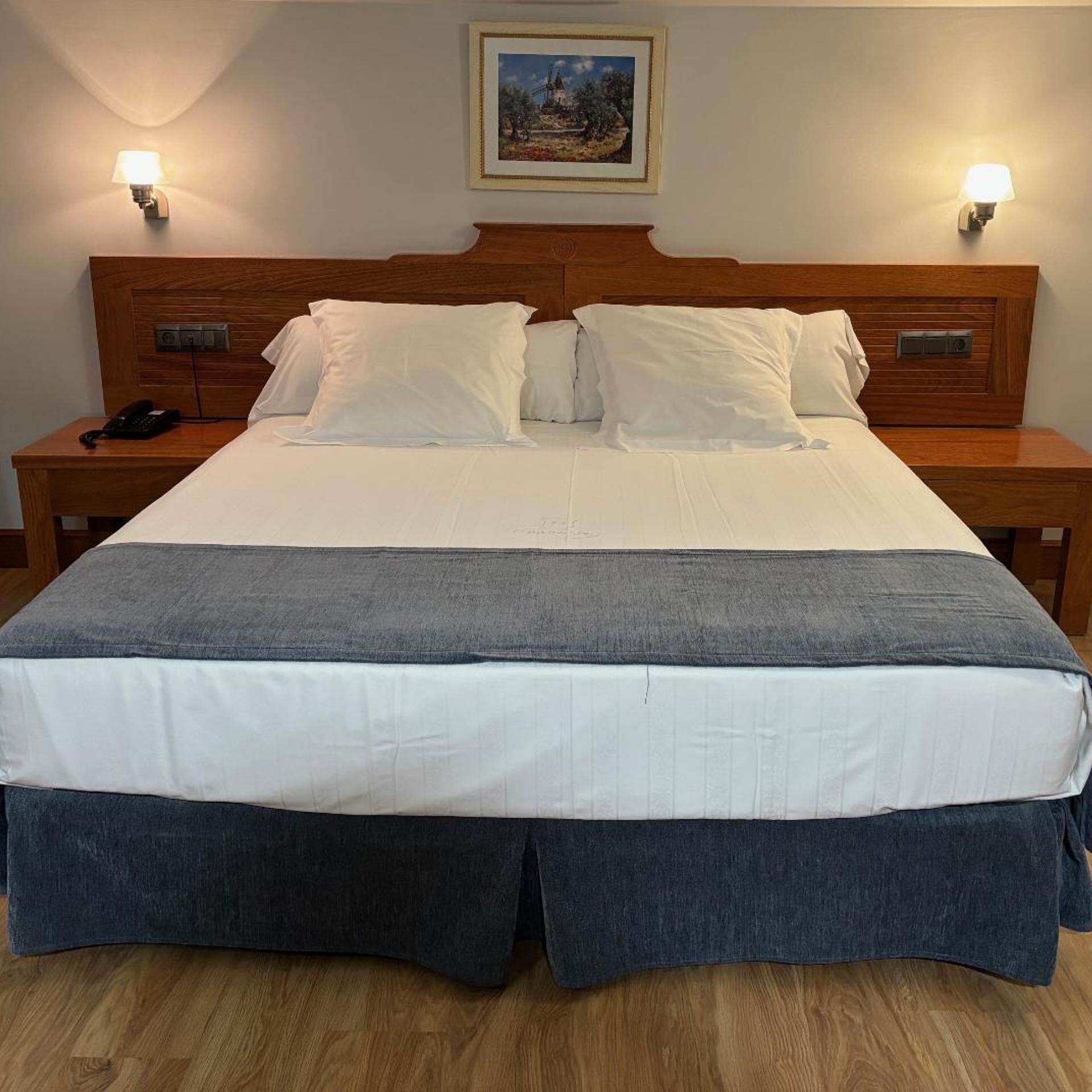 Una cama bien hecha con ropa de cama blanca y gris, dos mesitas de noche con lámparas y un cuadro en la pared.