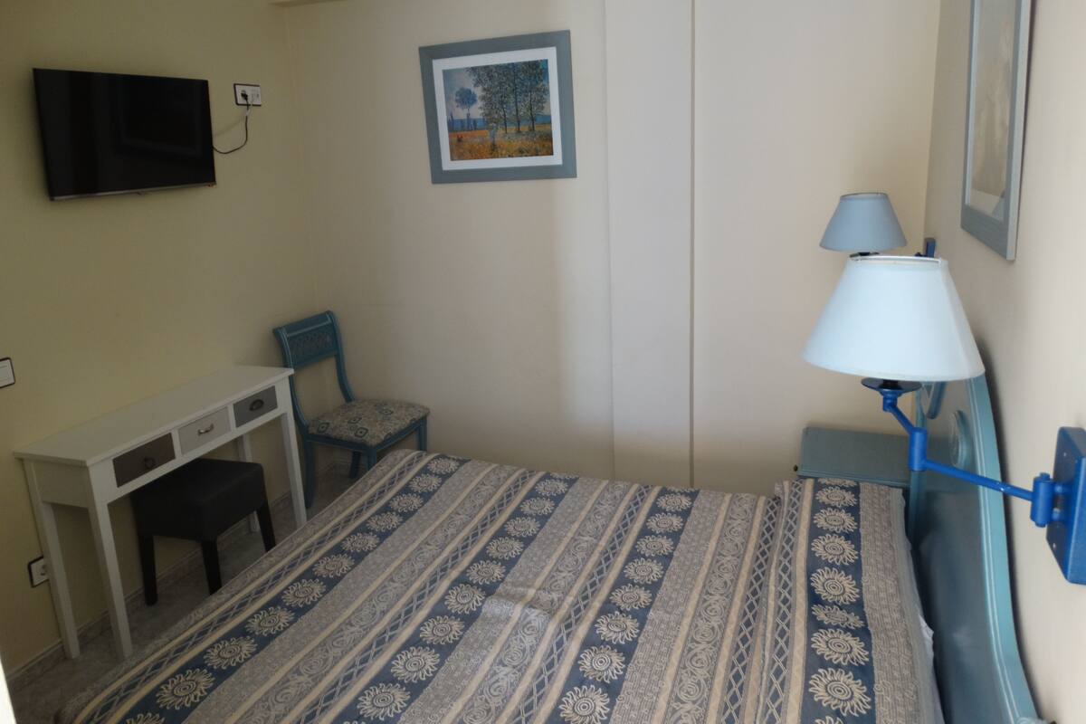 A room with a bed, a desk, a chair, a TV mounted on the wall, and a lamp.
