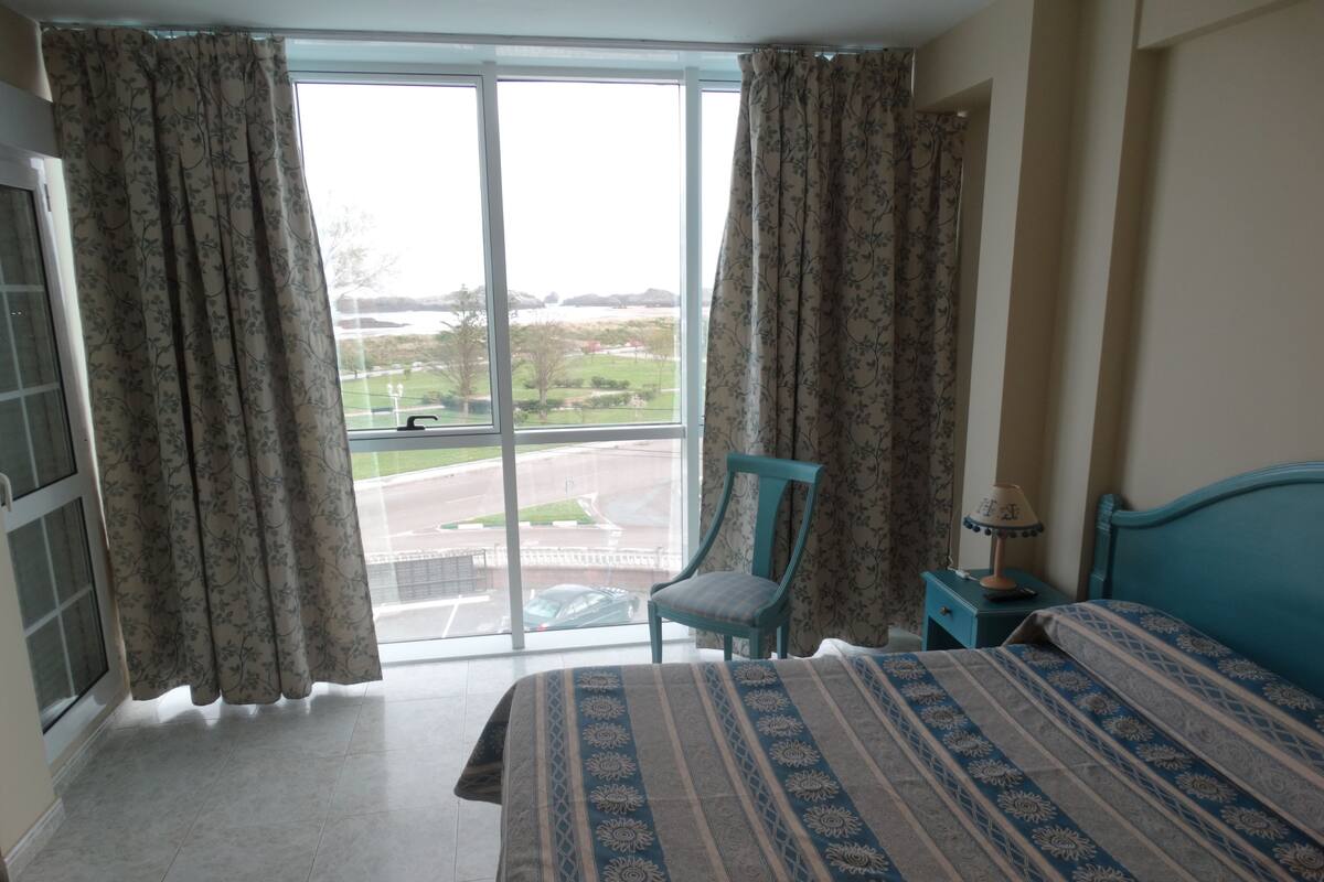 A bedroom with a bed, a chair, a nightstand, and a view of a road and greenery outside.