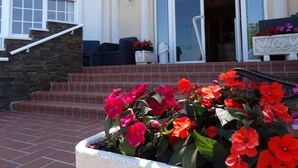 A building with a stone staircase, a door with glass panels, and a flower bed with red flowers. - Hotel Los Juncos (Noja)