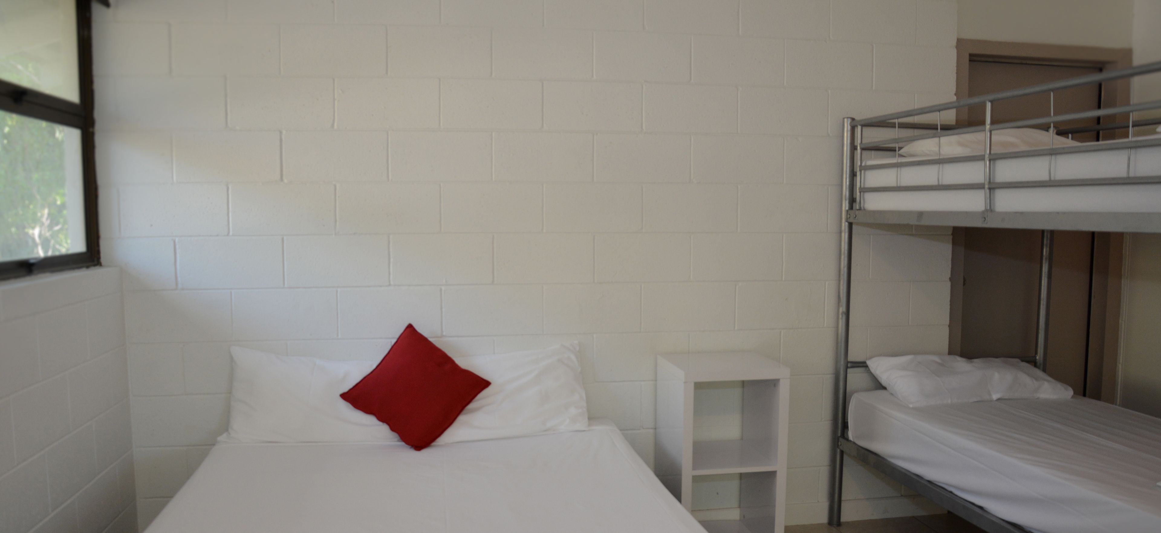 A single-bed room with white walls, a red pillow, and a white bedside table.