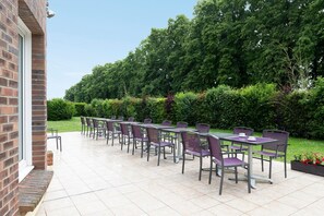 An outdoor patio with multiple tables and chairs, a brick building, and a well-maintained garden with trees and shrubs. - The Originals City, City Hôtel, Beauvais (Beauvais)