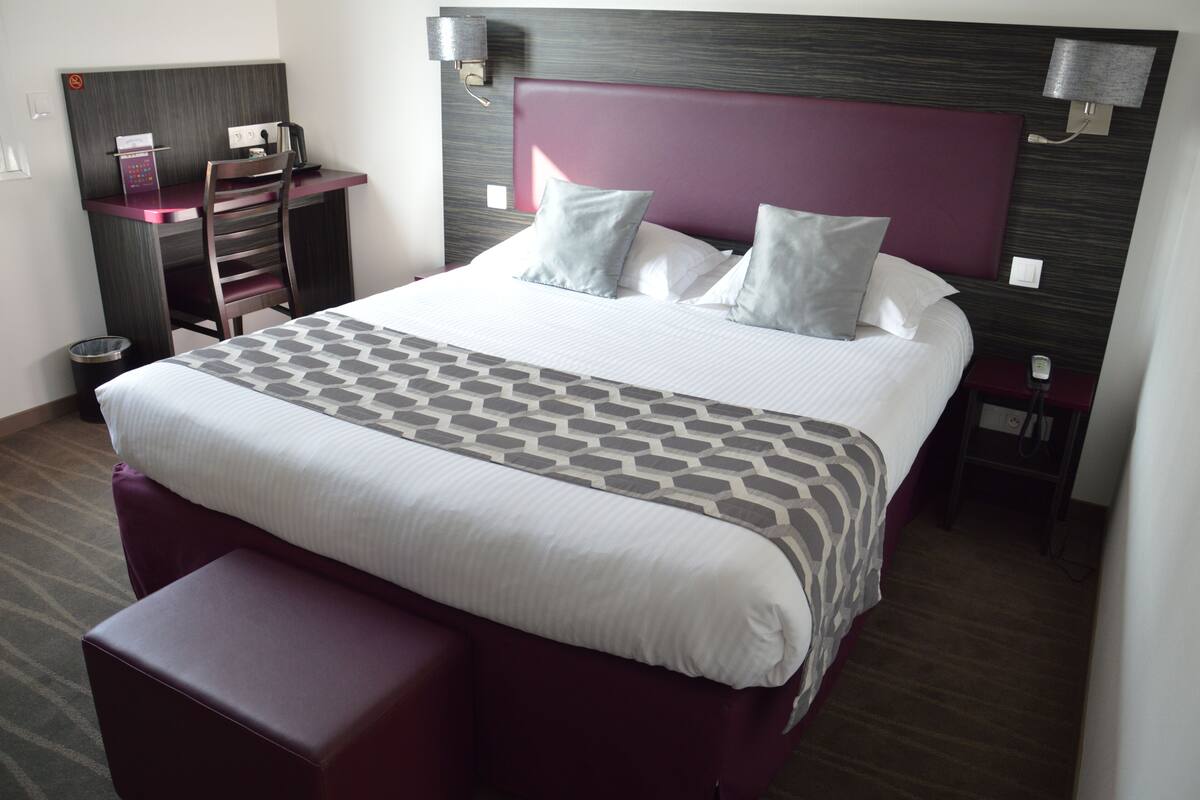 A neatly arranged hotel room with a large bed, a desk with a chair, and a small table.