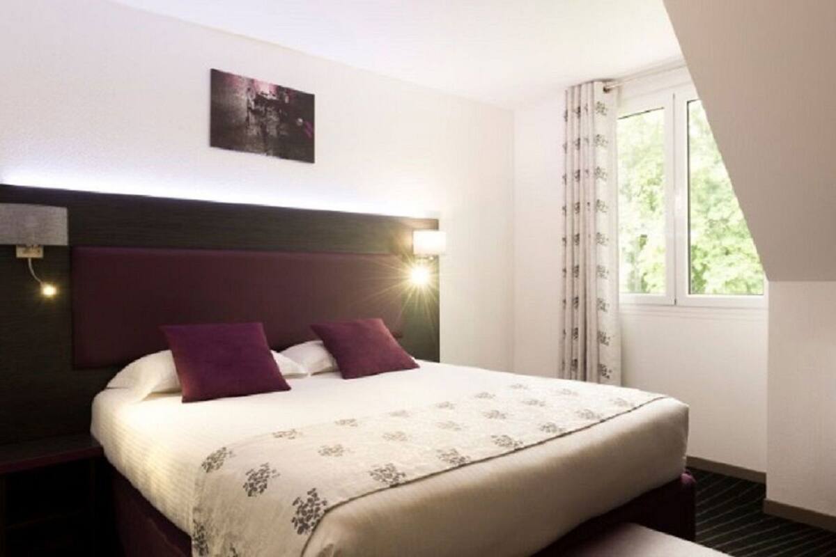 A hotel room with a bed, a headboard, a nightstand, a window with curtains, and a wall-mounted picture.