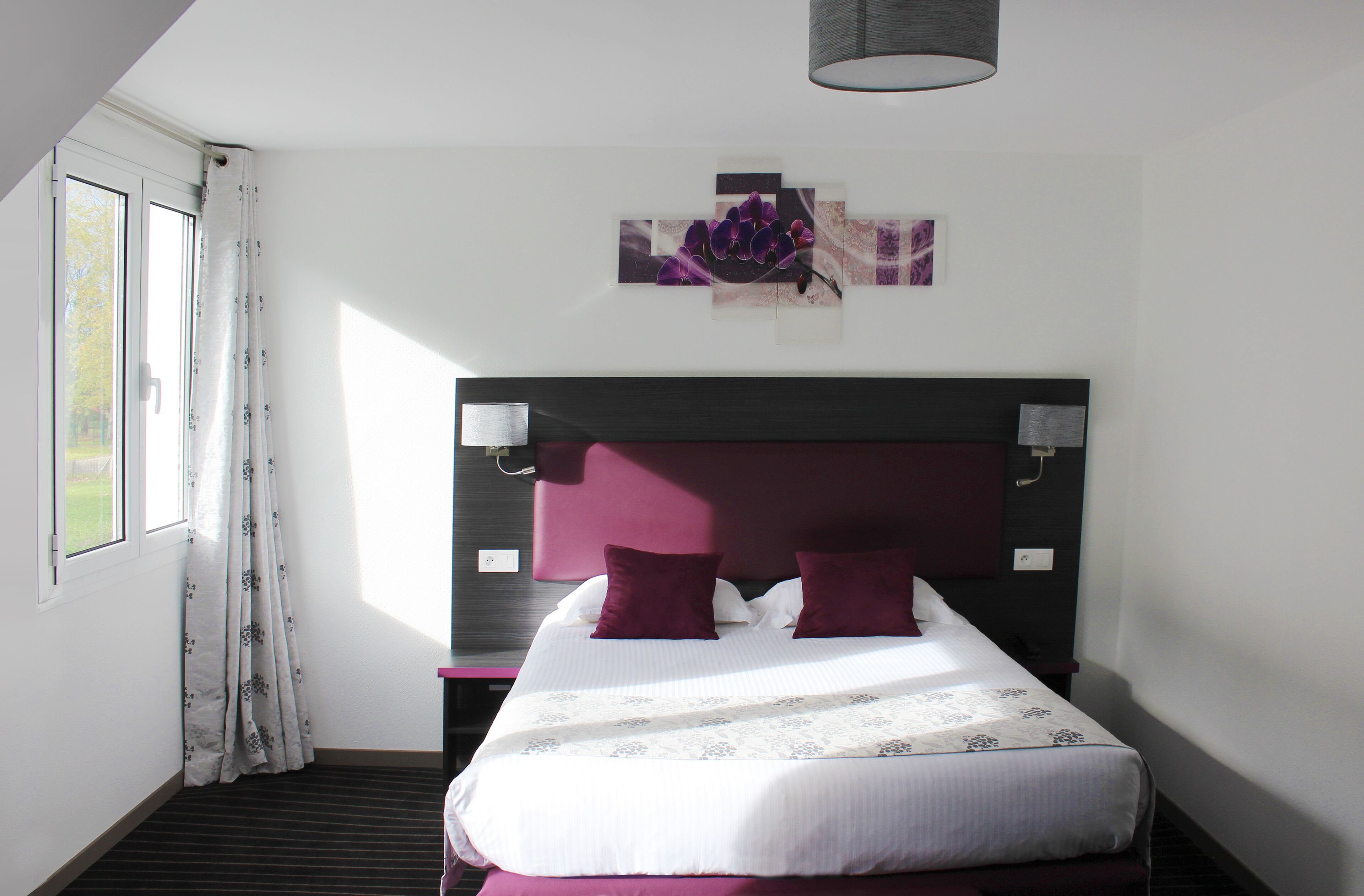 Une chambre d’hôtel avec un grand lit, une tête de lit, une table de chevet, une lampe et une fenêtre avec des rideaux.