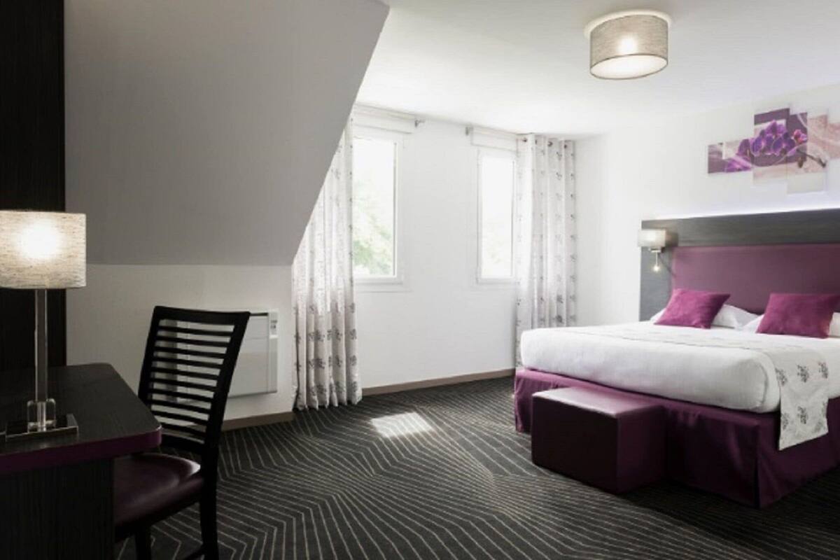 A hotel room with a bed, a desk, and a chair. There are windows with curtains, a lamp, and a wall-mounted artwork.