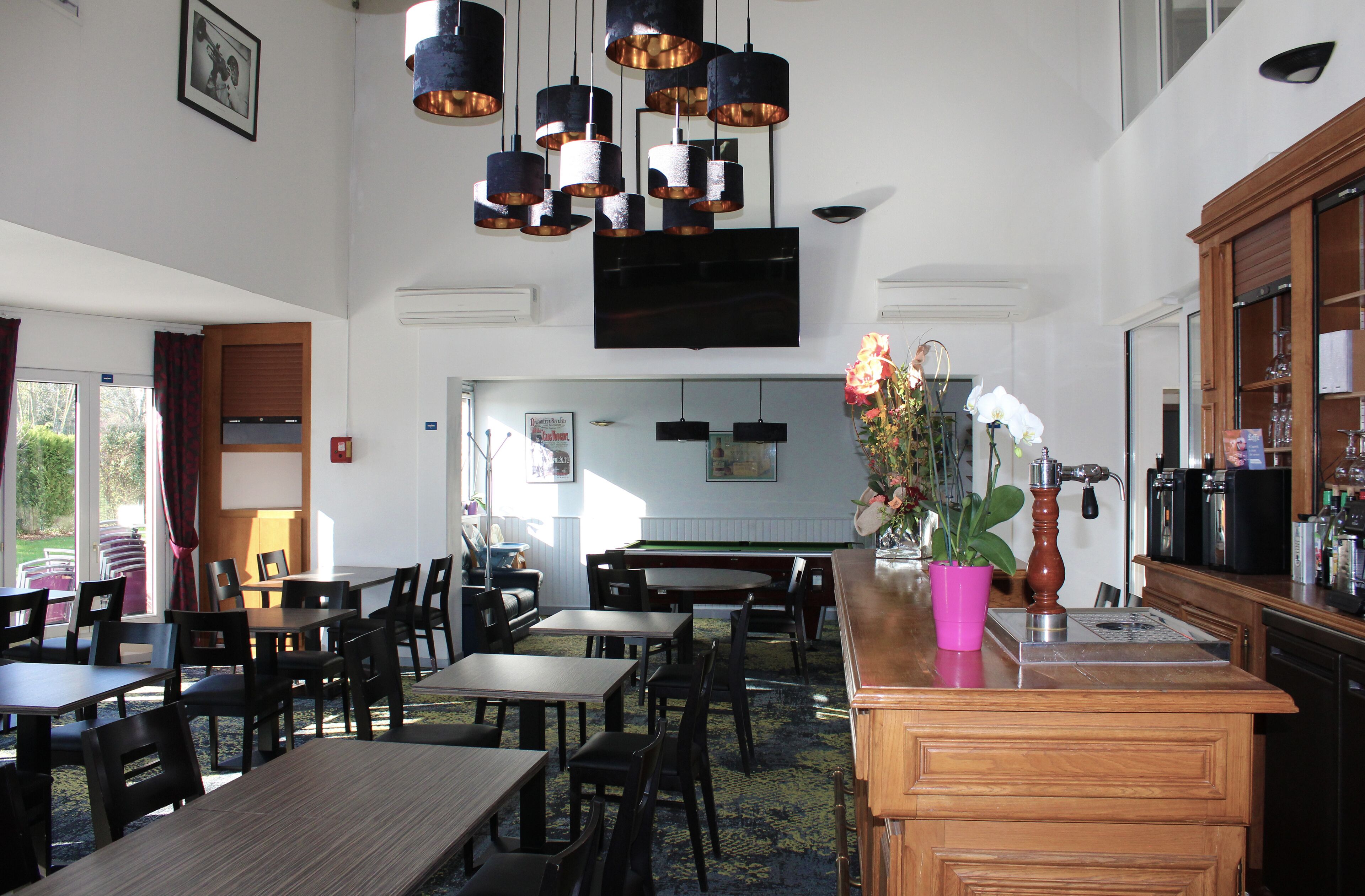 Un comedor amplio con varias mesas y sillas, una zona de bar con caja registradora, y un televisor de pantalla plana grande montado en la pared.