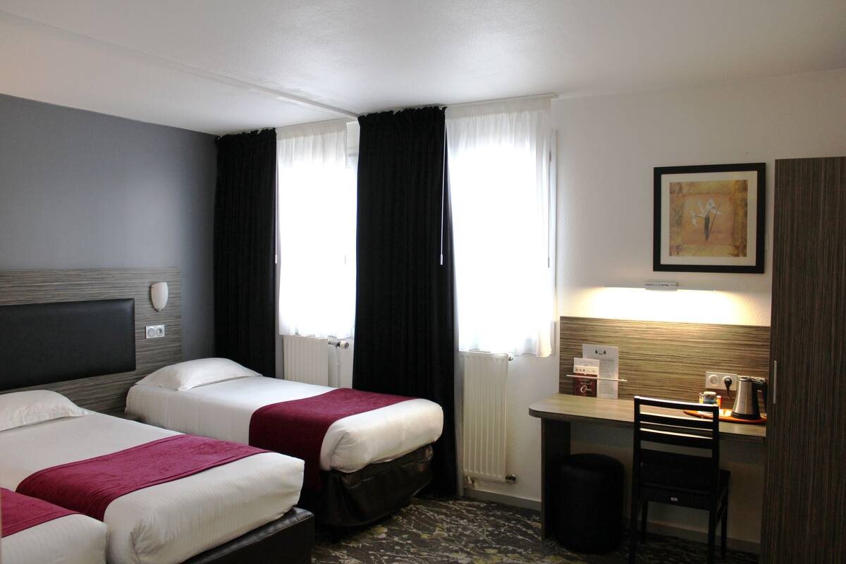 A hotel room with two beds, a desk, a chair, a radiator, and a framed picture on the wall.