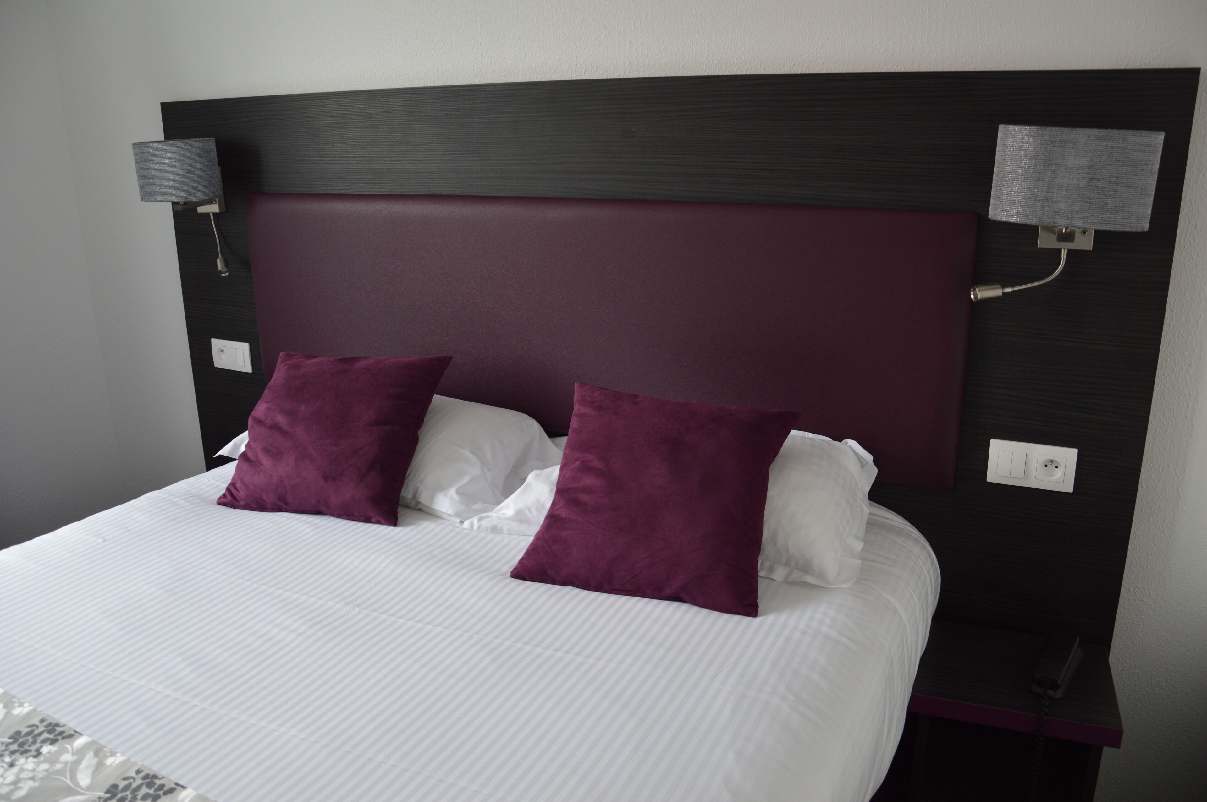 A neatly made bed with white linens and two purple pillows, a dark headboard, and two wall-mounted lamps.