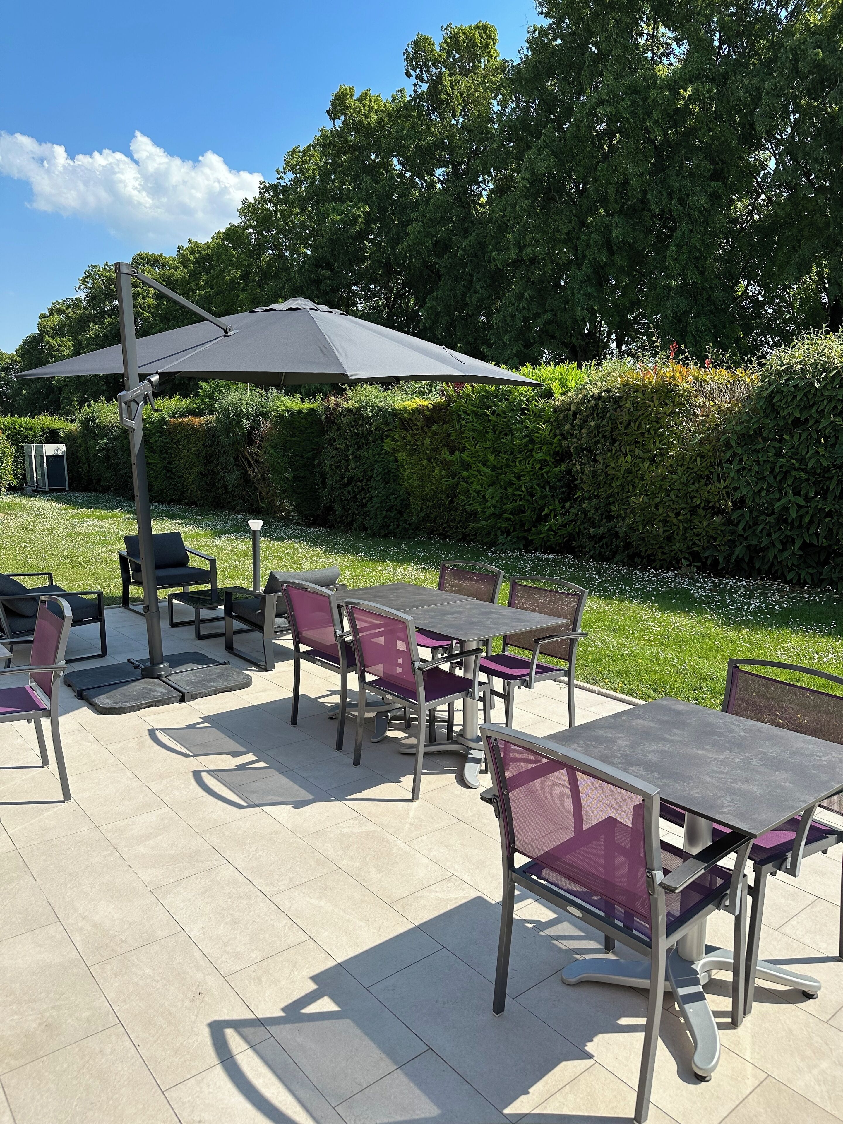 Un espace extérieur aménagé en terrasse, avec plusieurs tables et chaises, un grand parasol pour faire de l’ombre, et un jardin bien entretenu en arrière-plan.