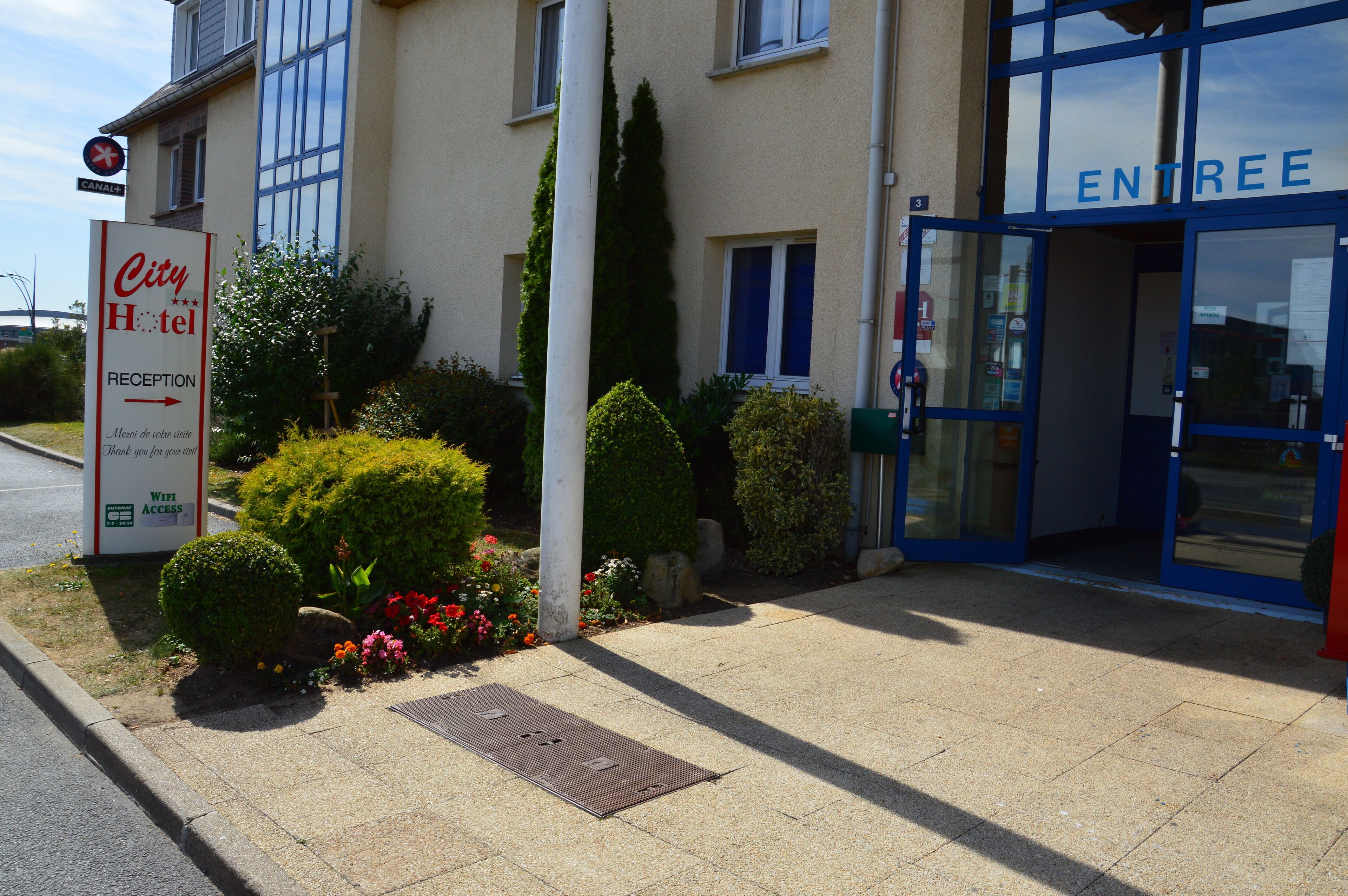 A hotel entrance with a reception sign, a blue door labeled "ENTREE", and a landscaped garden area.
