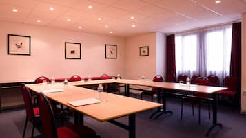 Uma sala de conferências com mesas compridas, cadeiras vermelhas, quadros nas paredes e uma janela grande com cortinas.