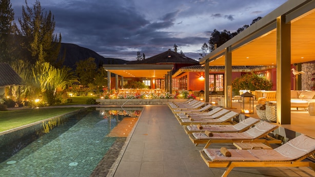 Zona de piscina con sillones de descanso y una piscina, rodeada por un jardín ajardinado y un edificio con porche cubierto.